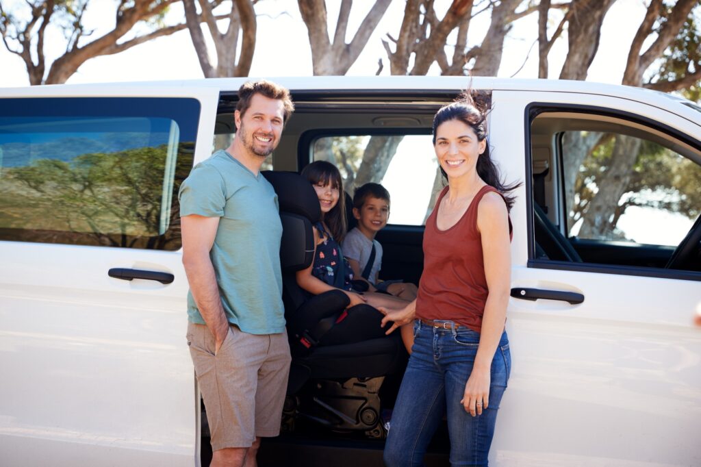 Mid adult white parents standing beside car, their two young kids inside smiling to camera